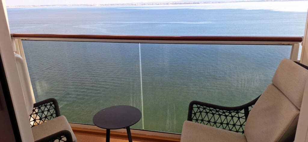 Vista al balcón con vista a una gran masa de agua. El balcón tiene una barandilla de cristal con barandilla de madera en la parte superior. Hay dos sillas con respaldo tejido y una pequeña mesa redonda entre ellas. El agua está tranquila y el horizonte se ve a lo lejos.