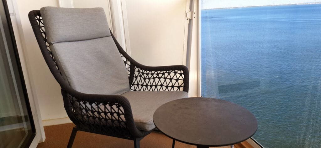 Un acogedor balcón con una silla de mimbre y una pequeña mesa redonda. El balcón da a una amplia masa de agua, proporcionando una vista tranquila y relajante.