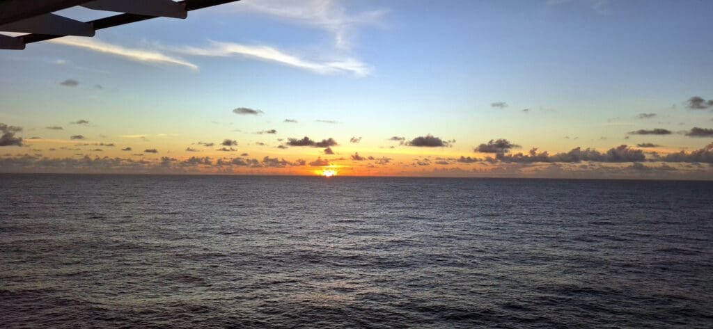 Tranquila vista al mar al atardecer, con el sol parcialmente visible en el horizonte. El cielo es de un tono azul y naranja, intercalado con pequeñas nubes. El océano parece en calma, reflejando los colores del atardecer. Parte de la estructura es visible en la esquina superior izquierda.