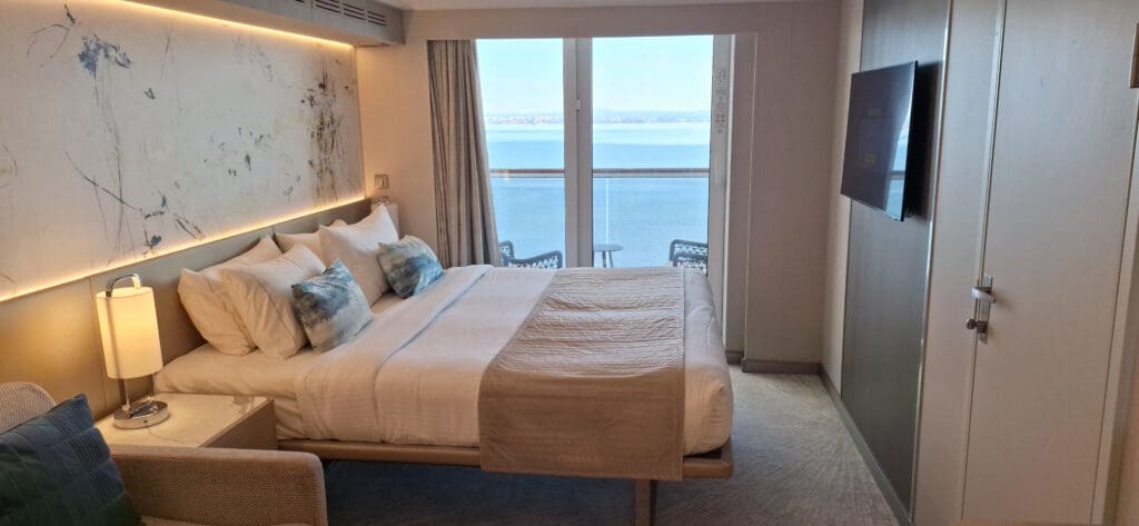 A modern hotel room with a large bed covered in white linens and several pillows. There is a nightstand with a lamp beside the bed. The room features a large abstract painting on the wall above the bed. A flat-screen TV is mounted on the opposite wall. The room has a large window with curtains, offering a view of the ocean and a distant shoreline.