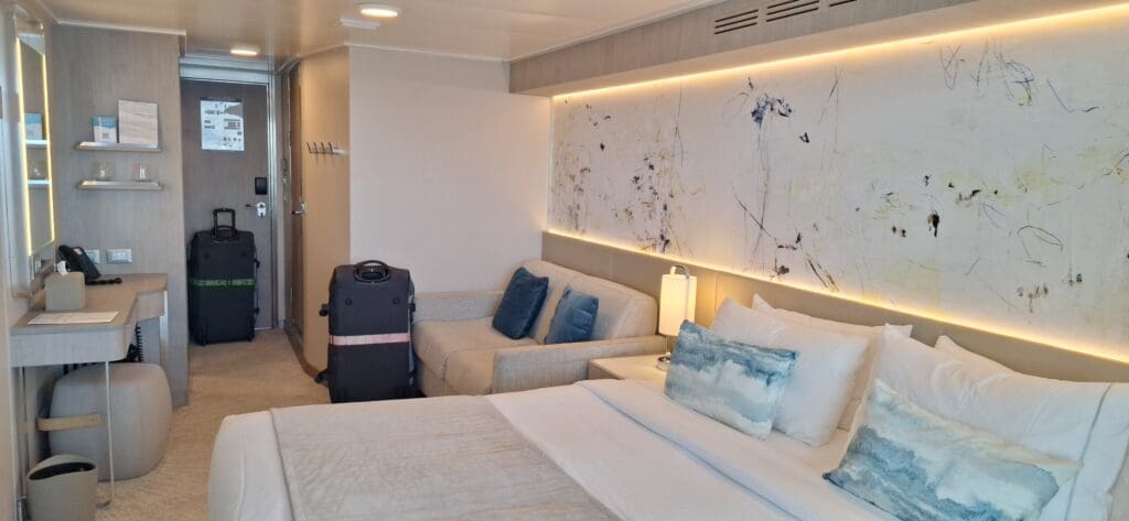 Una habitación de hotel moderna con una cama cuidadosamente hecha, sábanas blancas y almohadas azules. Al lado de la cama hay un sofá de color claro con cojines azules. Una gran obra de arte abstracta está colgada en la pared sobre el sofá, iluminada por una luz suave. Se colocan dos bolsas cerca de la puerta de entrada. Hay un pequeño escritorio con un teléfono y una silla en el lado izquierdo, y estantes con tazas y folletos montados en la pared. La habitación tiene una paleta de colores neutros con un aspecto limpio y organizado.