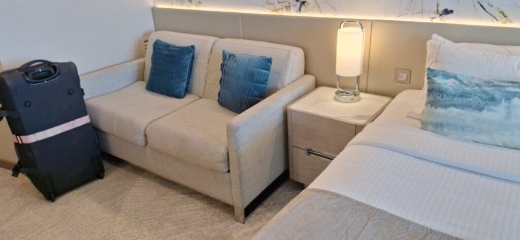 Una habitación de hotel con un sofá beige con dos almohadas azules, una maleta negra con un cinturón rosa al lado, una mesita de noche con una lámpara moderna y una elegante sección de cama con sábanas blancas y una almohada estampada azul.