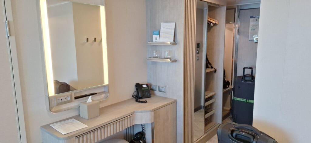 La foto muestra el interior de una habitación de hotel con un diseño moderno. Hay un espejo de pared con iluminación incorporada encima del escritorio. Sobre el escritorio hay un teléfono, una caja de pañuelos y algunos papeles. Encima del escritorio hay dos estantes con vasos y un pequeño cartel. A la derecha hay un armario abierto con perchas y estanterías. Delante del armario se ve parcialmente una maleta. La habitación tiene una paleta de colores neutros con madera clara y tonos beige.