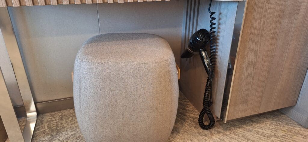 Se coloca una otomana tapizada en gris debajo de una mesa o escritorio de madera. A la derecha de la otomana, un secador de pelo negro con un cable enrollado está montado en el costado del tocador. El piso está alfombrado y el tocador tiene acabados en madera.