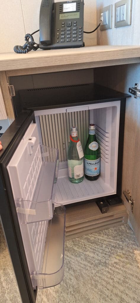 La foto muestra un mininevera abierta dentro de un mueble de madera. Dentro del frigorífico hay dos botellas de agua con gas. Encima del gabinete hay un teléfono de escritorio negro con un cable enrollado y al lado hay dos enchufes eléctricos.