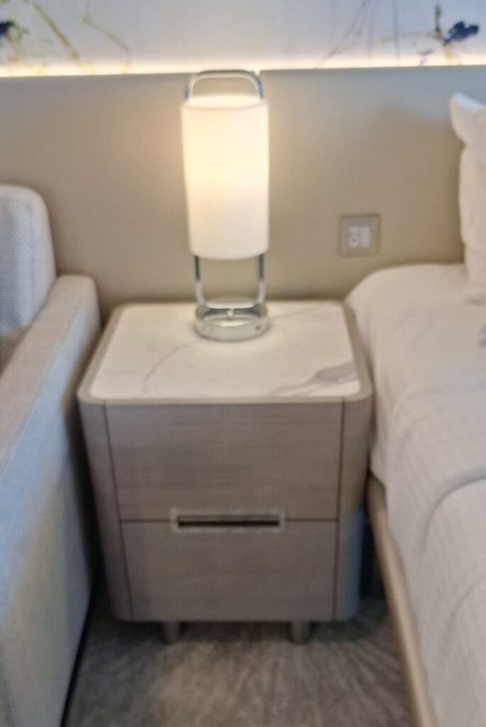 La foto muestra una mesita de noche con tapa de mármol, ubicada entre una cama y un sofá. Sobre la mesa hay una lámpara moderna con pantalla cilíndrica blanca y base de metal. La pared detrás de la mesa tiene un delicado patrón decorativo y en la pared se ve una toma de corriente.