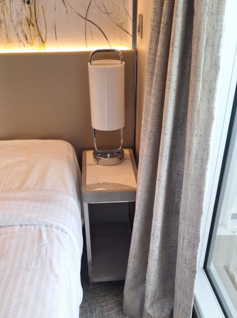 La foto muestra la esquina de un dormitorio moderno con una cama a la izquierda, cubierta con sábanas blancas. Al lado de la cama hay una pequeña mesita de noche con una elegante lámpara cilíndrica encima. La pared detrás de la cama presenta arte abstracto con iluminación tenue. A la derecha, unas cortinas que llegan hasta el suelo cubren parcialmente una ventana o una puerta de cristal.