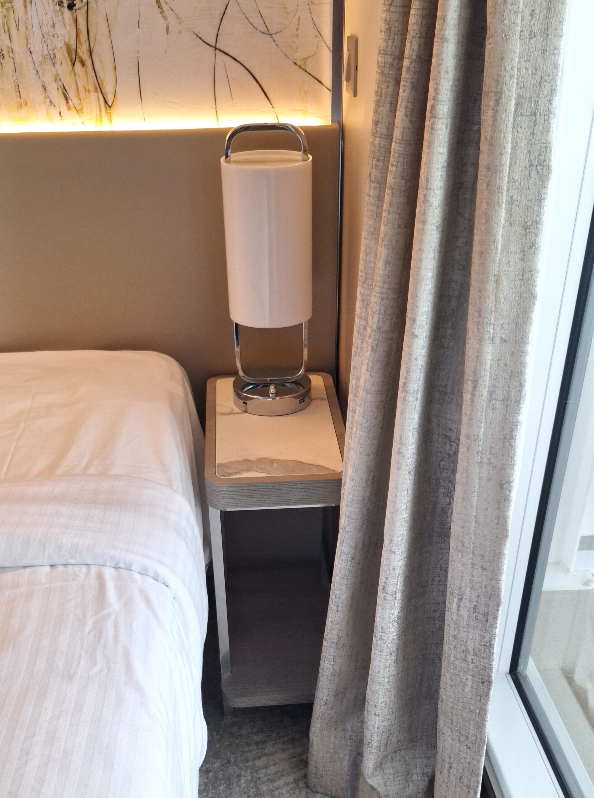 The image shows a modern bedroom corner with a bed on the left, covered in white sheets. Next to the bed is a small bedside table with a sleek, cylindrical lamp on top. The wall behind the bed has abstract art with subtle lighting. On the right, there are floor-length curtains partially covering a window or glass door.