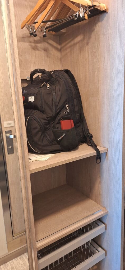 La foto muestra un mueble de madera abierto con varios colgadores de madera y metal sujetos a una barra en la parte superior. En el estante del medio hay una mochila negra con un objeto rojo en el bolsillo lateral. Debajo del estante hay tres cajones de rejilla.