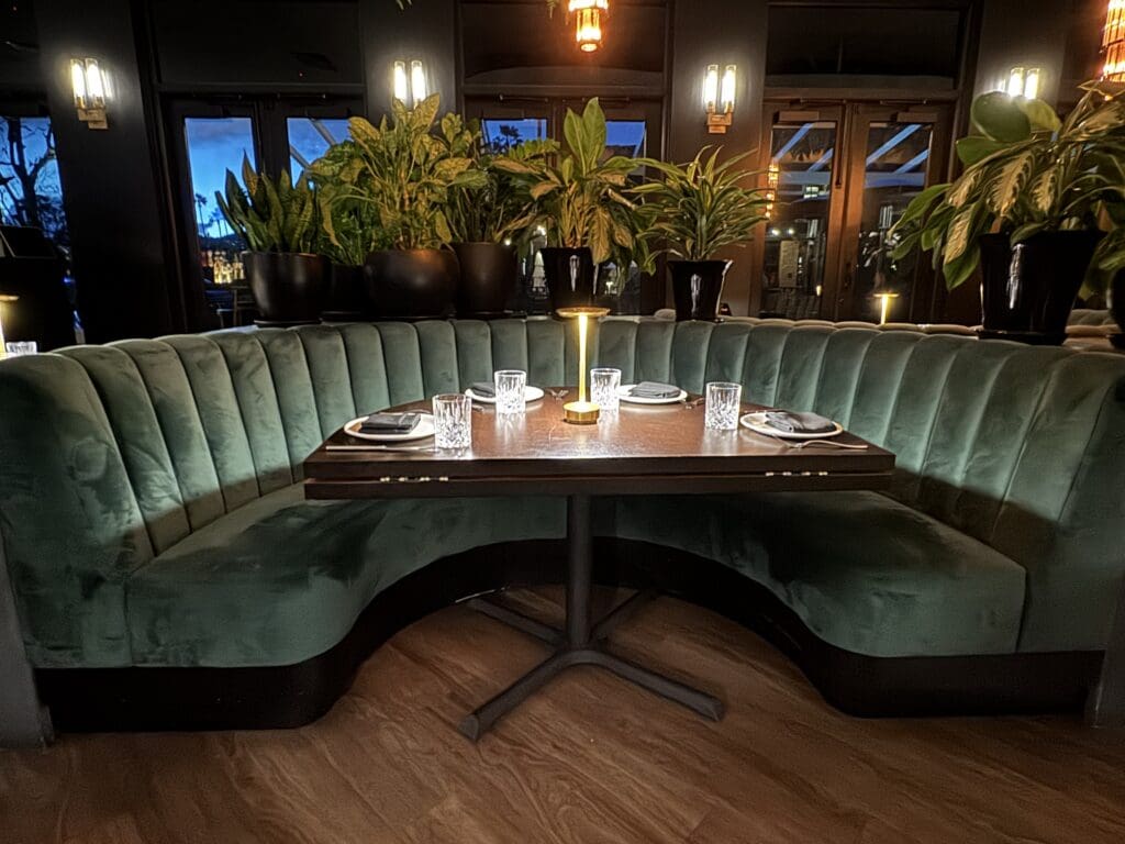 The image shows a cozy restaurant booth with a semi-circular green velvet sofa surrounding a dark wooden table. The table is set with plates, napkins, and glasses, and a small lamp provides warm lighting. Behind the booth, there are several large potted plants, and the background features windows with a view of the outside. The ambiance is elegant and inviting.
