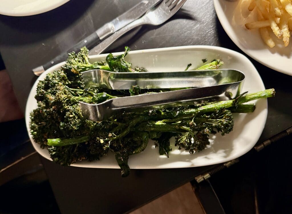 Broccolini at BLVD Steak in Calabasas, California. A white rectangular plate with cooked broccolini and metal tongs on top. The plate is on a dark table, with a fork and knife nearby. A portion of fries is visible on a separate plate in the background.