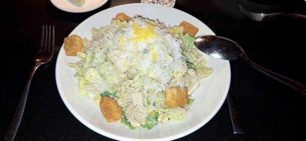 A Caesar salad is served in a white bowl, topped with grated cheese and croutons. A fork and spoon are placed on either side of the bowl, and a small plate with a lemon wedge is in the background.
