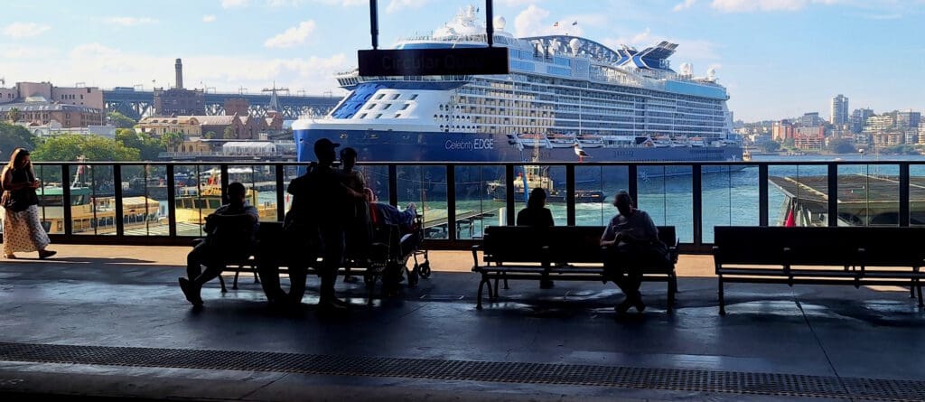 The image shows a large cruise ship, the "Celebrity Edge," docked at a harbor. In the foreground, there are silhouettes of people sitting and standing on benches at a terminal, with a sign above reading "Circular Quay." The background features a cityscape with buildings and a bridge, under a clear blue sky.