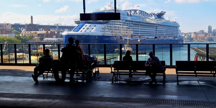 The image shows a large cruise ship, the "Celebrity Edge," docked at a harbor. In the foreground, there are silhouettes of people sitting and standing on benches at a terminal, with a sign above reading "Circular Quay." The background features a cityscape with buildings and a bridge, under a clear blue sky.