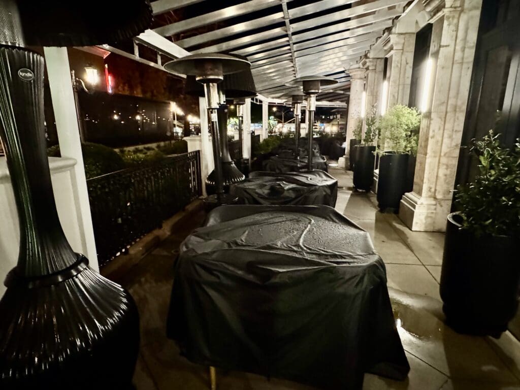 Patio at BLVD Steak in Calabasas, California. The image shows an outdoor patio area with tables covered in dark protective covers. There are several tall outdoor heaters positioned between the tables. The area is partially enclosed with a transparent roof and surrounded by potted plants and decorative columns. The scene is dimly lit, suggesting it is nighttime.