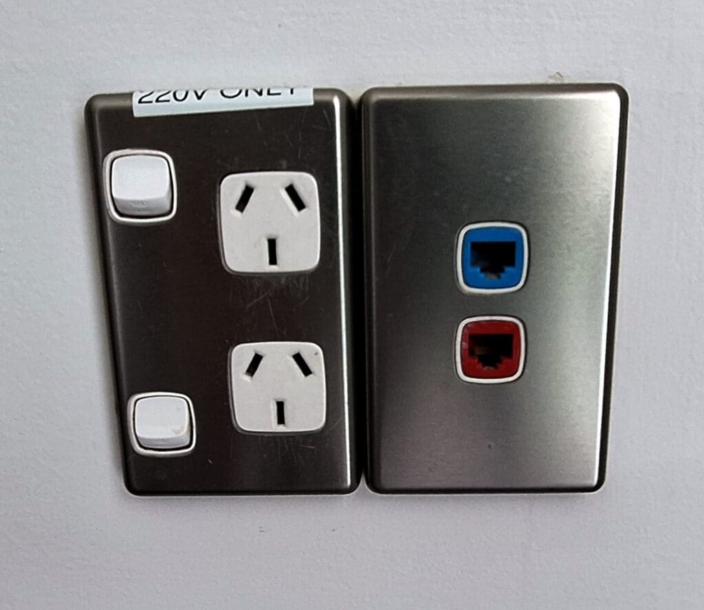 The image shows two wall plates. The left plate has two white electrical outlets with switches, labeled "220V ONLY." The right plate has two network ports, one with a blue border and the other with a red border. Both plates are mounted on a white wall.