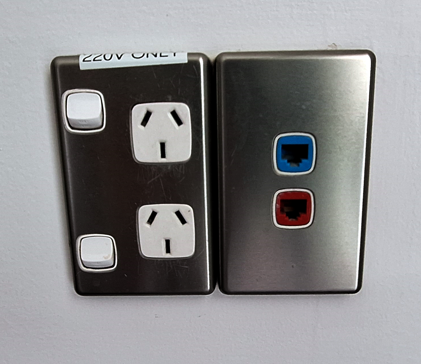 The image shows two wall plates. The left plate has two white electrical outlets with switches, labeled "220V ONLY." The right plate has two network ports, one with a blue border and the other with a red border. Both plates are mounted on a white wall.