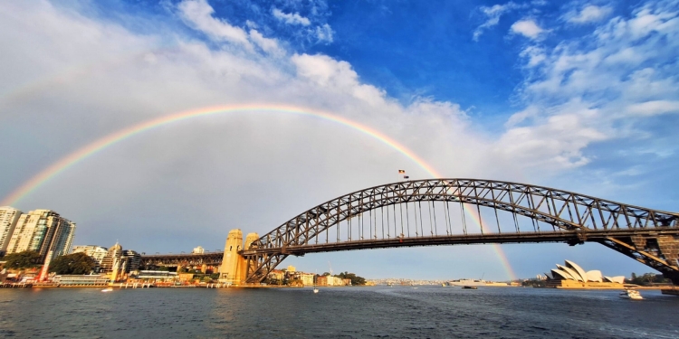 A panoramic view of Sydney Harbour featuring the Sydney Harbour Bridge and the Sydney Opera House. A vibrant rainbow arches across the sky, with a partly cloudy blue sky in the background. The water in the foreground reflects the scene, and buildings are visible along the shoreline.