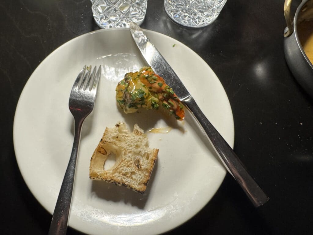 A white plate on a dark table holds a piece of bread and a small portion of food garnished with herbs. A fork and knife are placed on the plate. In the background, there are two glasses with a textured pattern.