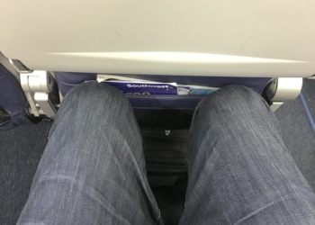 Tight seat reclining on an airline. The image shows a view from an airplane seat, focusing on the limited legroom. A person's legs, wearing blue jeans, are visible, with knees close to the seat in front. The seatback pocket contains a magazine or safety card. The overall setting is inside an airplane cabin.