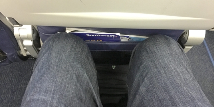 Tight seat reclining on an airline. The image shows a view from an airplane seat, focusing on the limited legroom. A person's legs, wearing blue jeans, are visible, with knees close to the seat in front. The seatback pocket contains a magazine or safety card. The overall setting is inside an airplane cabin.