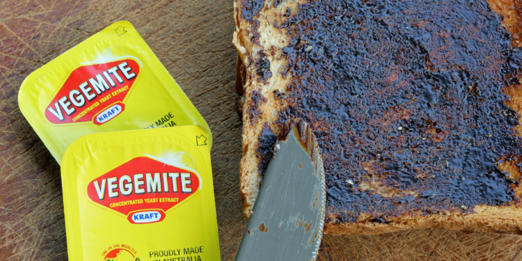The image shows two small yellow packets of Vegemite, a concentrated yeast extract, placed on a wooden surface. Next to them is a slice of bread spread with Vegemite, and a knife with some Vegemite on its blade rests on the bread.