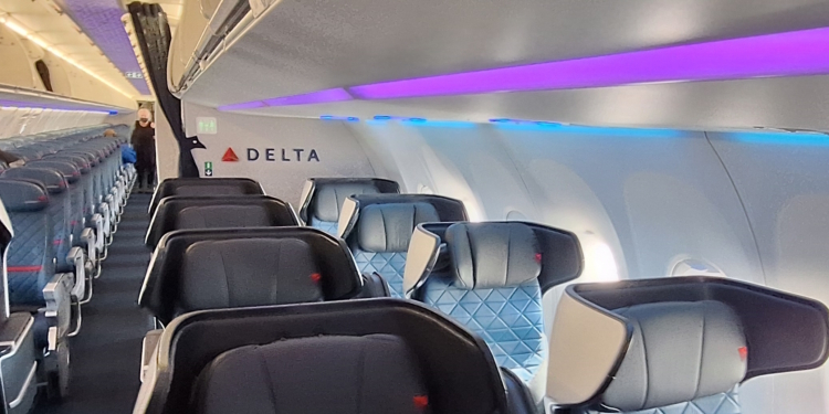 The image shows the interior of an airplane cabin, specifically the first-class section. The seats are wide and have high backs with headrests, upholstered in dark material with a quilted pattern. The cabin lighting is a mix of purple and blue, creating a modern ambiance. The word "DELTA" is visible on the wall, indicating the airline. The aisle leads to the economy section in the background, where more seats are visible.
