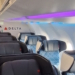 The image shows the interior of an airplane cabin, specifically the first-class section. The seats are wide and have high backs with headrests, upholstered in dark material with a quilted pattern. The cabin lighting is a mix of purple and blue, creating a modern ambiance. The word "DELTA" is visible on the wall, indicating the airline. The aisle leads to the economy section in the background, where more seats are visible.