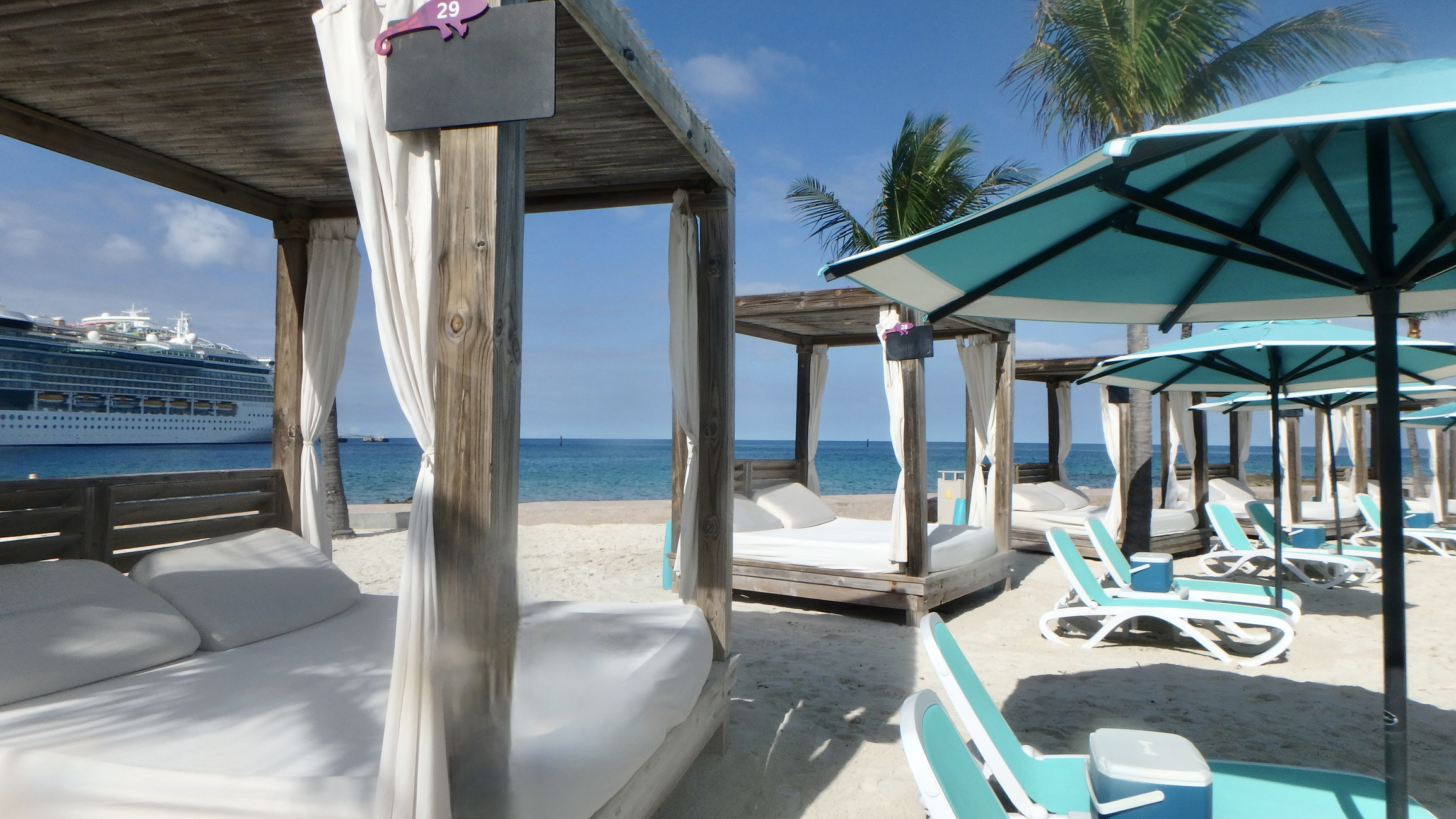 Daybeds and loungers on Chill Island Beach on Royal Caribbean Cruise line's private island. The image shows a beach scene with several cabanas and lounge chairs under teal umbrellas on a sandy shore. In the background, there is a large cruise ship on the ocean. Palm trees are visible, and the sky is clear and blue.