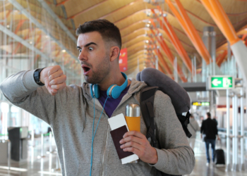 A man in an airport looks surprised as he checks his watch. He is wearing a gray hoodie, blue headphones around his neck, and has a backpack with a neck pillow attached. He holds a passport, a boarding pass, and a travel mug. The background shows the airport's modern architecture and a green exit sign.