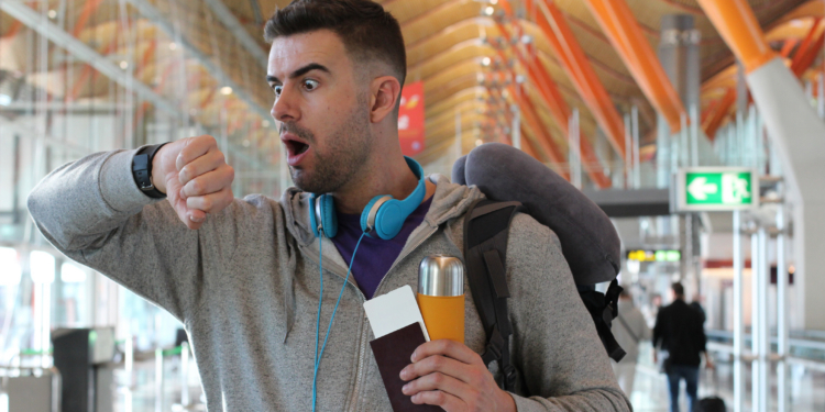 A man in an airport looks surprised as he checks his watch. He is wearing a gray hoodie, blue headphones around his neck, and has a backpack with a neck pillow attached. He holds a passport, a boarding pass, and a travel mug. The background shows the airport's modern architecture and a green exit sign.