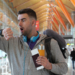A man in an airport looks surprised as he checks his watch. He is wearing a gray hoodie, blue headphones around his neck, and has a backpack with a neck pillow attached. He holds a passport, a boarding pass, and a travel mug. The background shows the airport's modern architecture and a green exit sign.