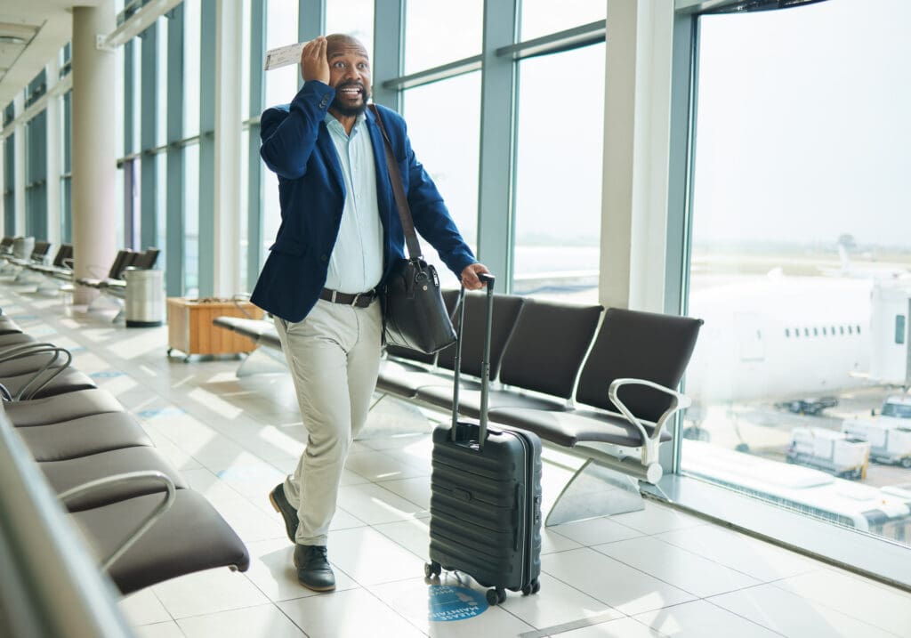 A man is walking through an airport terminal, looking concerned. He is holding a boarding pass to his forehead and pulling a black suitcase with one hand. He is wearing a blue blazer, light blue shirt, beige pants, and black shoes. The terminal has large windows with a view of an airplane outside. There are empty seats along the side of the walkway.