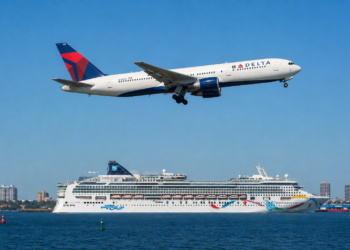 A Delta Air Lines 767 flies over an NCL cruise ship in a harbor