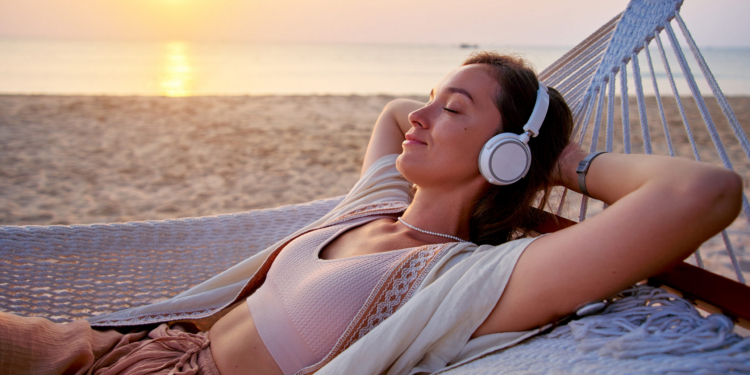 Young blissful calm woman wearing wireless headphones with closed eyes and hands behind head lie in a hammock and enjoy relaxing music