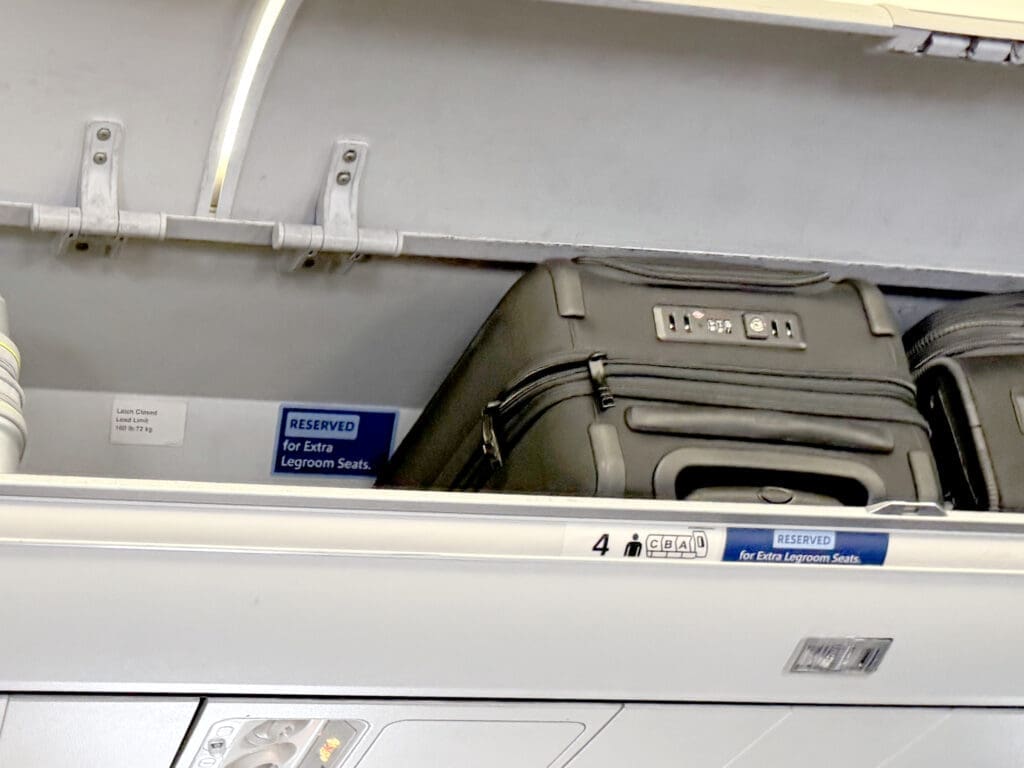 Overhead bin with a blue "Reserved for Extra Legroom Seats" placard and a hard-shell roller bag stowed above row 4