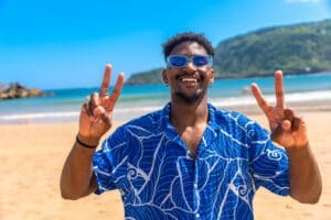 Young tourist enjoying summer holidays on a tropical beach, wearing sunglasses and a blue shirt, flashing a peace sign