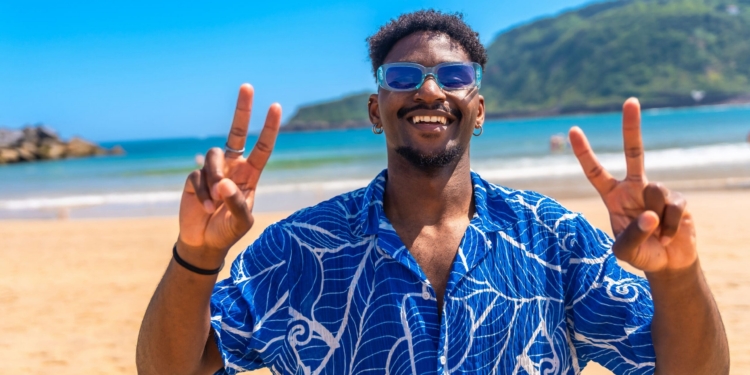 Young tourist enjoying summer holidays on a tropical beach, wearing sunglasses and a blue shirt, flashing a peace sign