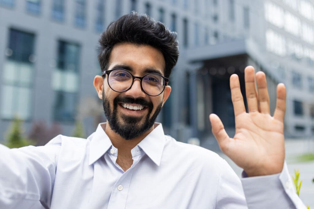 Young successful businessman outside office building walking in daytime, man smiling waving hand to phone camera, worker smiling contentedly, using video call app on phone