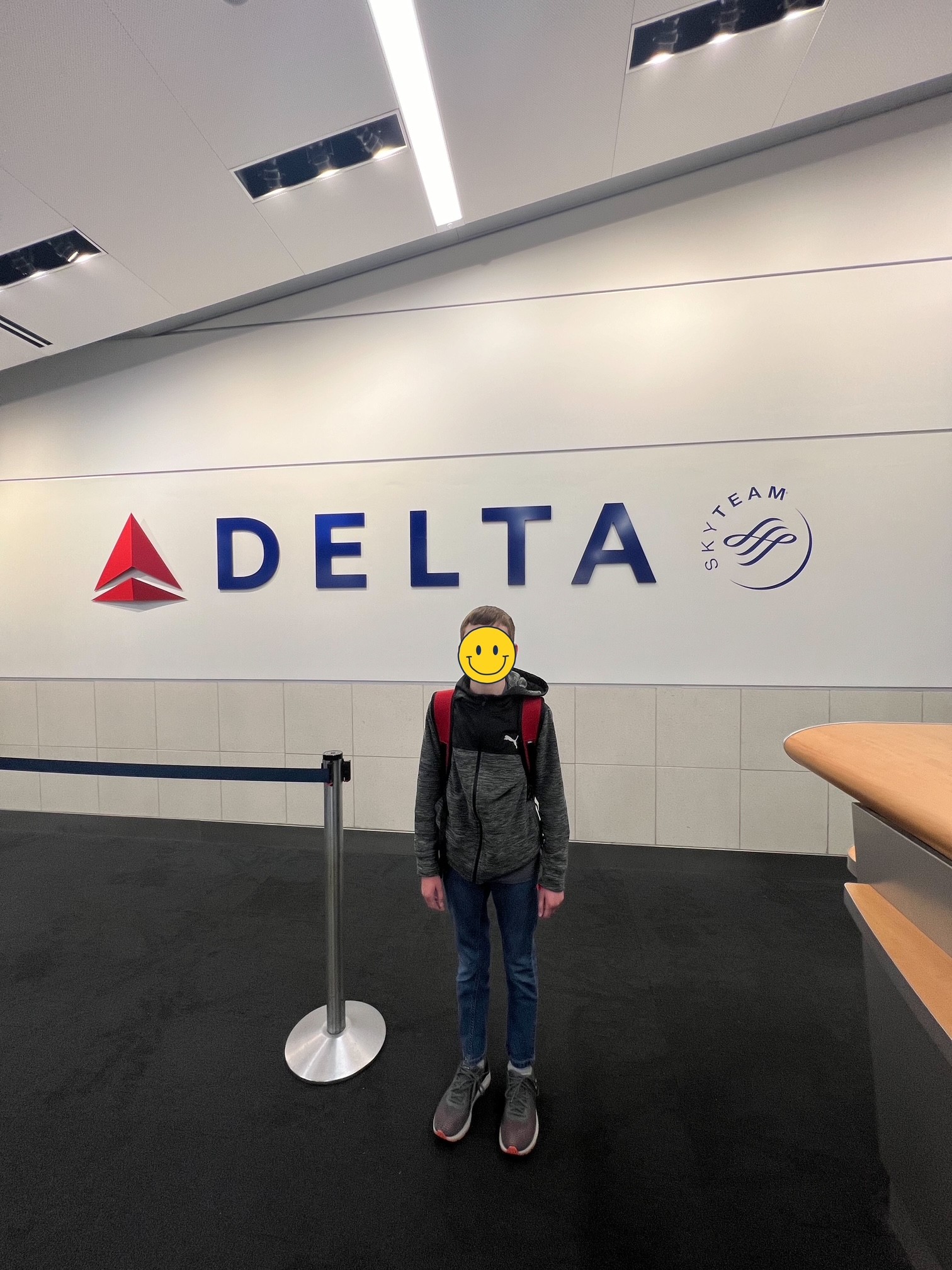 A person with a smiley face sticker covering their face is standing in front of a wall with the Delta Air Lines logo and the SkyTeam logo. The person is wearing a gray jacket, blue jeans, gray sneakers, and a red backpack. There is a stanchion with a blue belt next to them, and part of a counter is visible on the right.