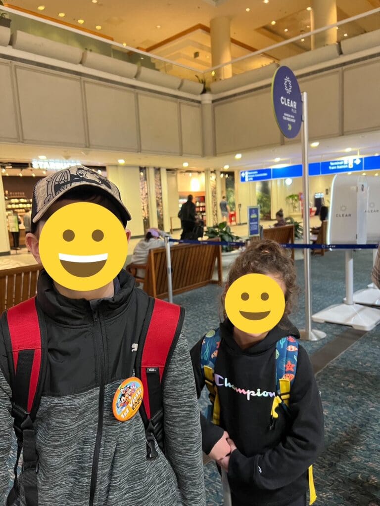 Two children are standing indoors, both wearing backpacks. The child on the left is wearing a cap and a jacket with a badge, while the child on the right is wearing a hoodie. They are in a public area with signs and seating visible in the background.