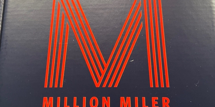 The image shows a dark blue box with the words "MILLION MILER" in red capital letters. Above the text is a large red letter "M" made of parallel lines. In the top right corner, there is a red and blue triangular logo. The box is placed on a speckled surface.