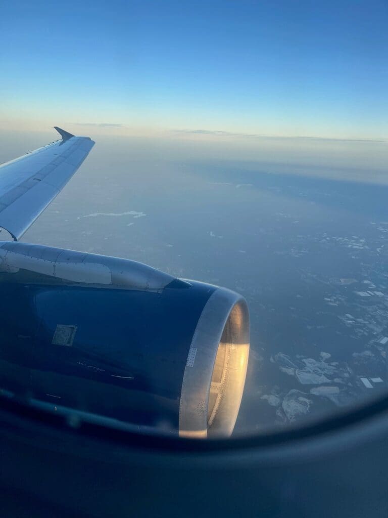 The image shows a view from an airplane window, capturing part of the aircraft's wing and engine. The sky is clear with a gradient from light to deep blue, and the landscape below is partially visible, showing patches of land and water. The sunlight is reflecting off the engine.