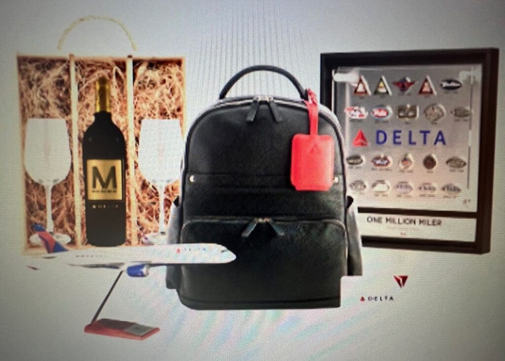 The image shows a collection of items including a black backpack with a red tag, a framed display with various logos and the word "DELTA," a model airplane, a bottle of wine labeled "M" in a wooden box with two wine glasses, and a small card with the Delta logo. The framed display also has the text "ONE MILLION MILER."