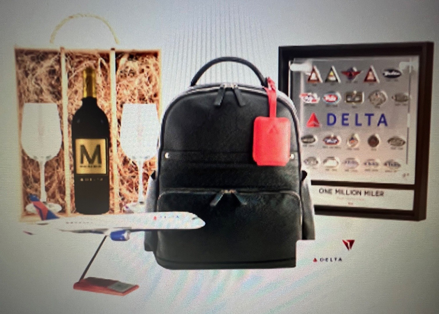 The image shows a collection of items including a black backpack with a red tag, a framed display with various logos and the word "DELTA," a model airplane, a bottle of wine labeled "M" in a wooden box with two wine glasses, and a small card with the Delta logo. The framed display also has the text "ONE MILLION MILER."