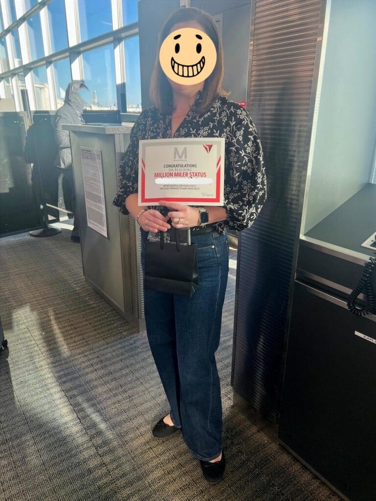 A person is standing indoors, holding a certificate that reads "Congratulations on reaching Million Miler Status." They are wearing a black and white patterned blouse and blue jeans. Their face is covered with a smiley face graphic. The background shows large windows and another person in a hooded jacket.