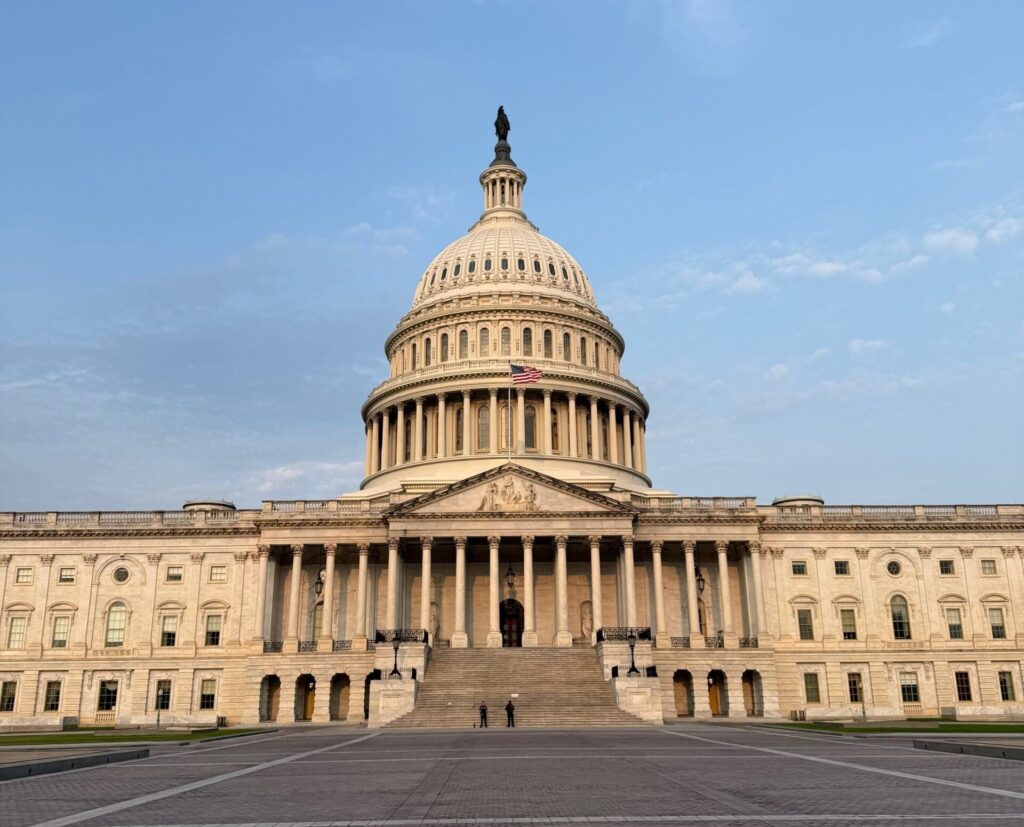 The image shows the United States Capitol building in Washington, D.C. It features a large dome at the center, flanked by two wings. The building is made of white stone and has a series of columns at the front entrance. The sky is clear and blue.